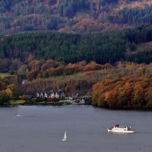 A large white ferry and a sailboat on a lake with a shoreline of buildings and autumn-colored forests.