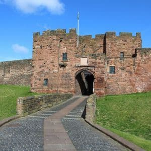A cobblestone path leads over a bridge to the gatehouse of a large red stone castle under a clear blue sky.