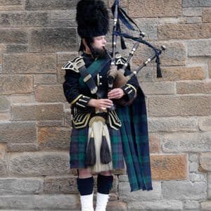 A man in traditional Scottish dress, including a tartan kilt and tall bearskin hat, plays the bagpipes in front of a stone wall.