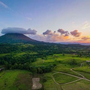 Una vista aerea di un vulcano con la sua cima tra le nuvole, che domina una valle verde lussureggiante e una piccola città al tramonto.