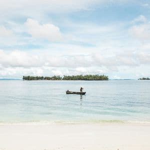 Una persona pagaia una piccola canoa su acque calme al largo di una spiaggia di sabbia bianca, con un'isola ricoperta di palme all'orizzonte.