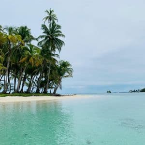 Una piccola isola tropicale con una spiaggia di sabbia bianca e molte palme, circondata da acque turchesi e cristalline sotto un cielo nuvoloso.