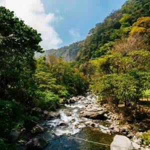 Un fiume roccioso scorre attraverso una valle circondata da montagne lussureggianti e boscose sotto un cielo azzurro parzialmente nuvoloso.