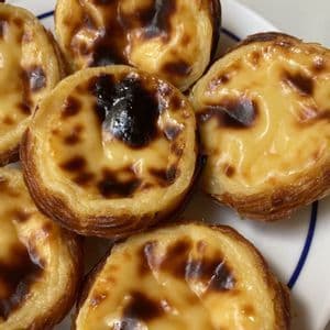 Un primo piano di alcuni pasteis de nata con croste friabili e la superficie caramellata su un piatto bianco.