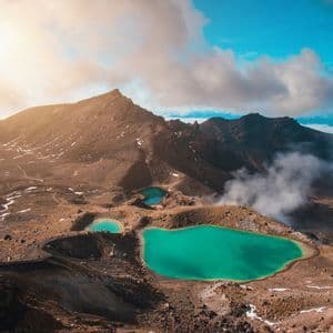An aerial view of three turquoise crater lakes nestled in a rocky, volcanic landscape with steam rising from the ground.