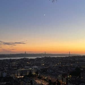 Un paesaggio urbano al crepuscolo visto dall'alto, con edifici illuminati, un ponte distante sull'acqua e una luna crescente nel cielo colorato.