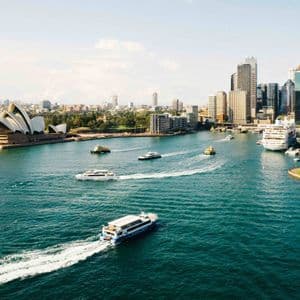 An aerial view of a busy harbor with ferries, a large opera house, and a modern city skyline under a clear sky.