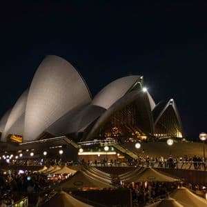 The Sydney Opera House illuminated at night, viewed from a crowded waterfront promenade with outdoor seating.