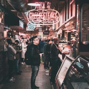 People waiting in line at an indoor food market under a large red 'Pizza & Cheesesteaks' neon sign.