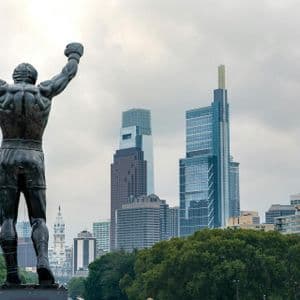 The back of a bronze statue of a boxer with raised arms in a victory pose, overlooking a modern city skyline under a cloudy sky.