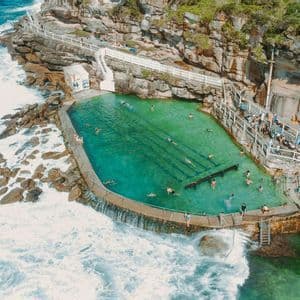 An aerial view of an ocean pool carved into a rocky cliff, with people swimming as waves crash against the barrier.
