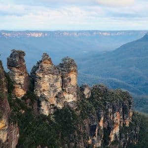 Three large rock pillars stand on a cliff edge overlooking a wide, forested mountain valley under a cloudy sky.