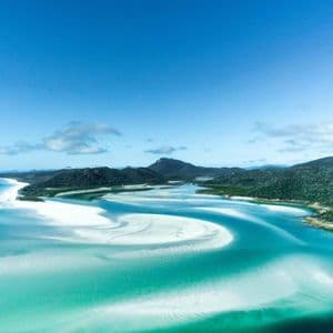 An aerial view of turquoise water swirling around white sandbars and green, hilly islands under a clear blue sky.