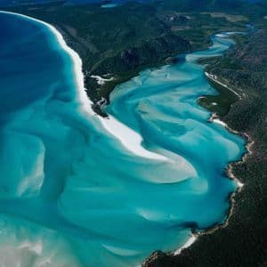 An aerial view of white sand swirling through vibrant turquoise water along a forested coastline, with distant islands on the horizon.