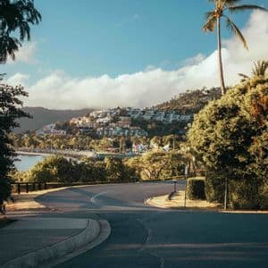 A curving road with palm trees overlooks a seaside town built into a green hillside under a partly cloudy sky.