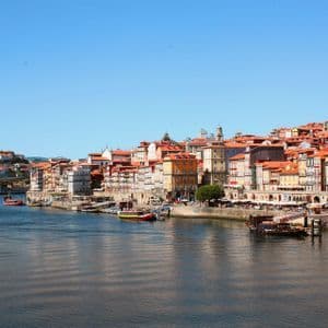 Edifici colorati su un pendio costeggiano un fiume, con barche ormeggiate lungo la passeggiata sul lungofiume sotto un cielo azzurro chiaro.