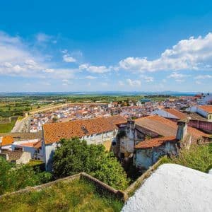 Veduta dall'alto di una cittadina con tetti di tegole rosse ed edifici bianchi, adagiata su un paesaggio verde sotto un cielo azzurro nuvoloso.