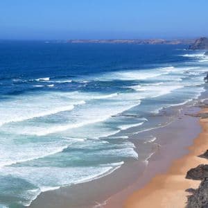 Onde bianche dell'oceano si infrangono su una spiaggia sabbiosa accanto a scogliere ripide e rocciose sotto un cielo azzurro e limpido.