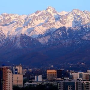 Eine Stadtlandschaft mit Gebäuden liegt am Fuße eines weitläufigen, schneebedeckten Gebirges, das von Sonnenlicht unter klarem Himmel beleuchtet wird.