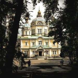 Eine gelbe orthodoxe Kathedrale mit farbenfrohen Kuppeln und goldenen Spitzen, aus der Ferne durch einen Rahmen dunkler Bäume betrachtet.