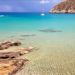 Una barca a vela bianca galleggia in una baia con acqua limpida e turchese, una costa rocciosa in primo piano e una costa collinare in lontananza.