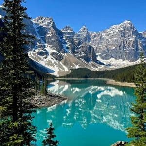 A vibrant turquoise lake reflects a range of snow-capped mountains, surrounded by a dense evergreen forest.