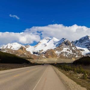 A paved road runs through a valley towards a range of towering, snow-capped mountains under a bright blue sky.