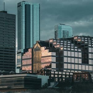 A cityscape of modern skyscrapers with glass facades reflecting the sunset under a dark, cloudy sky.