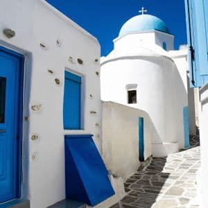 Un angusto vicolo in pietra tra edifici imbiancati a calce con porte blu, che conduce a una chiesa dalla cupola azzurra sotto un cielo limpido.