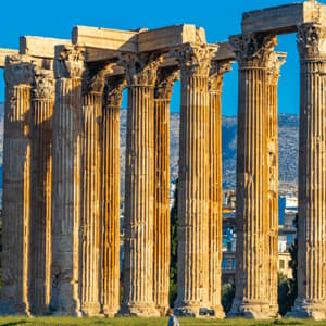 Alte colonne corinzie baciate dal sole di un'antica rovina, con una città e montagne visibili sullo sfondo sotto un cielo blu.