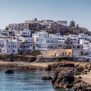 Un borgo collinare di case bianche si affaccia su un mare calmo, con persone che passeggiano lungo un molo di pietra.