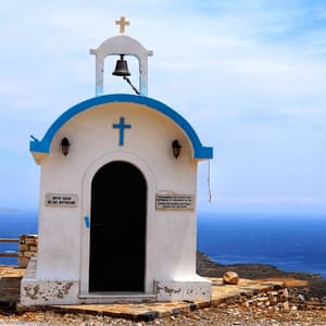 Una piccola cappella imbiancata, con dettagli blu e un campanile, si erge su una scogliera a picco sul mare azzurro.