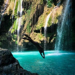 A person dives headfirst from a rock into a turquoise pool of water at the base of lush, cascading waterfalls.