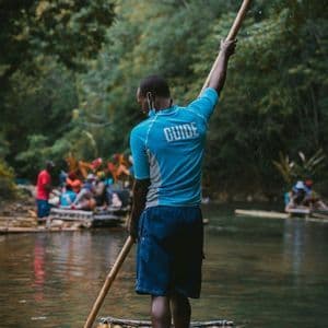 A guide seen from behind stands on a bamboo raft, using a long pole to navigate a river with a WeRoad group trip in the background.