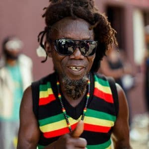 A man with dreadlocks and sunglasses smiles and gives a thumbs-up while wearing a striped vest.