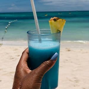 A hand with blue nail polish holds a frosty blue cocktail with a pineapple slice on a white sand beach.