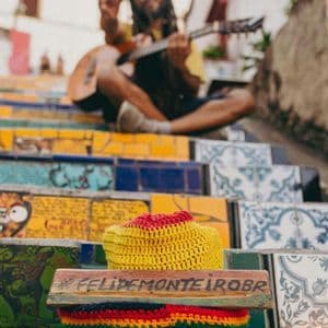 A man with dreadlocks plays guitar and makes a peace sign while sitting on colorful, tiled mosaic stairs, with a hat in the foreground.