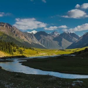 Ein Fluss schlängelt sich durch ein üppig grünes Tal auf eine entfernte Kette schneebedeckter Berge zu, wo eine WeRoad-Gruppe am Ufer rastet.
