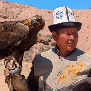 Ein Mann in traditioneller Kleidung hält einen Steinadler auf seiner behandschuhten Hand in einer hügeligen, trockenen Landschaft unter einem klaren blauen Himmel.