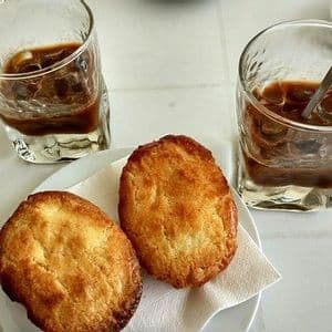 Due bicchieri di caffè freddo e due pasticcini rotondi su un piatto posati su un tavolo bianco.