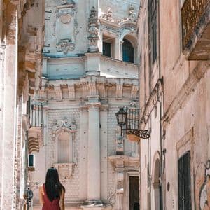 Una donna in tuta rossa vista di spalle, in piedi in uno stretto vicolo di pietra che guarda un ornato edificio barocco.