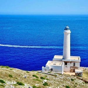 Un faro bianco si erge su una scogliera rocciosa a picco su un mare blu profondo, mentre un motoscafo sfreccia.