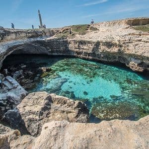 Una piscina naturale di acqua cristallina turchese all'interno di una caletta rocciosa, con persone che camminano sulle scogliere soprastanti sotto un cielo azzurro.