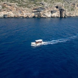 Una veduta aerea di un viaggio di gruppo WeRoad su una barca bianca che naviga in acque blu profonde verso una scogliera rocciosa con grotte marine.