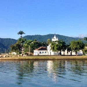 Eine weiße Kolonialkirche an einer üppigen Küste spiegelt sich im Wasser, mit grünen Hügeln im Hintergrund unter klarem Himmel.