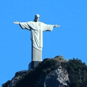Eine imposante Männerstatue ragt mit ausgebreiteten Armen auf einem felsigen Berggipfel unter klarem blauem Himmel empor.