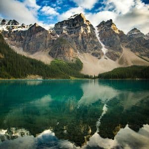 A rocky, snow-capped mountain range reflects onto the calm, turquoise water of a lake, with a pine forest on the shore.