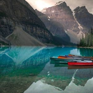 A row of colorful canoes docked on a pier next to a turquoise lake reflecting large, snow-capped mountains.