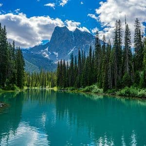 A calm turquoise lake reflects a pine forest and a jagged mountain peak with patches of snow under a blue, partly cloudy sky.