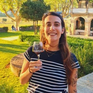 Una donna sorridente con un tatuaggio tiene in mano un bicchiere di vino rosso mentre si trova in un giardino soleggiato con alberi.